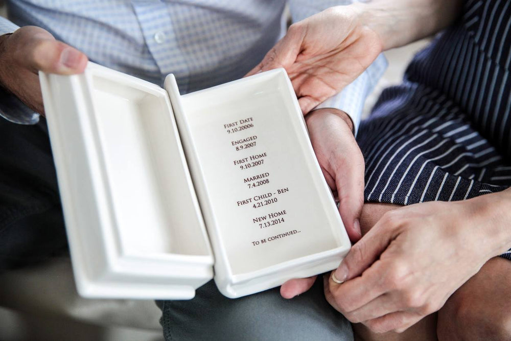 A white ceramic keepsake book box with a custom text label. The box is being held by a person with a blurred background.