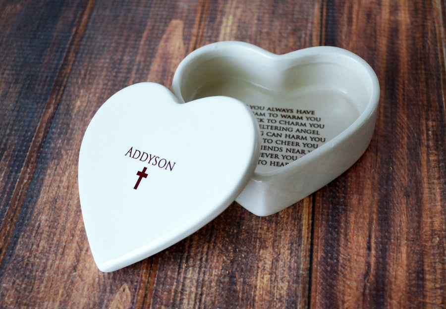 A white heart-shaped earthenware keepsake box with a child's name printed on the lid, featuring a cross image, and an open lid showing the interior with text.