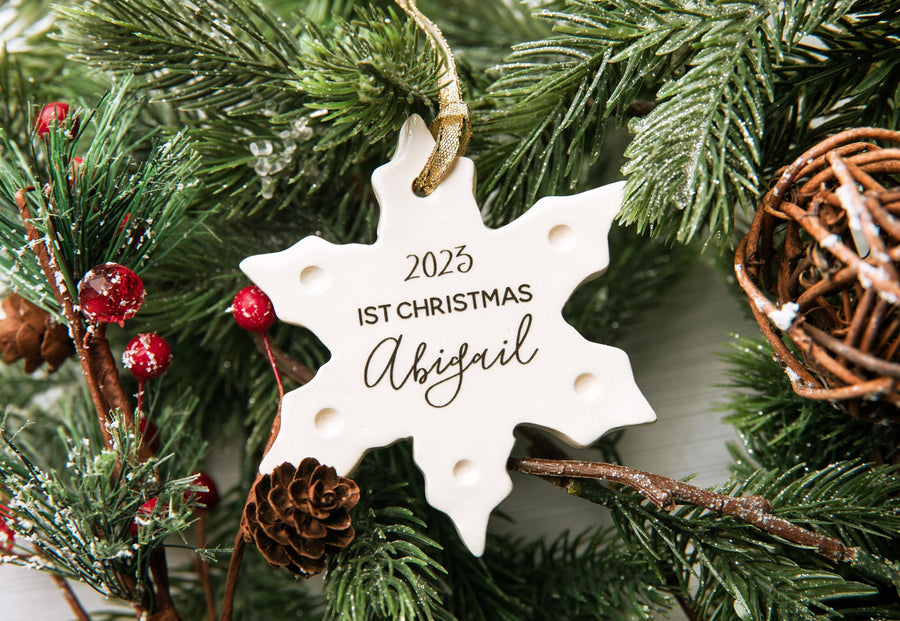A personalized ceramic snowflake ornament with the text '1st Christmas Abigail 2025' written on it, hanging on a Christmas tree.