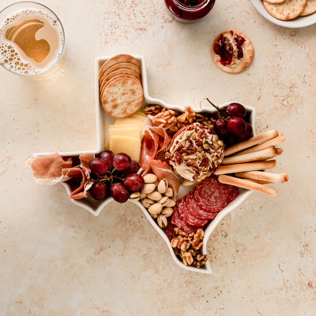 Texas-shaped hish with charcuterie with various meats, cheeses, and snacks on a light surface.