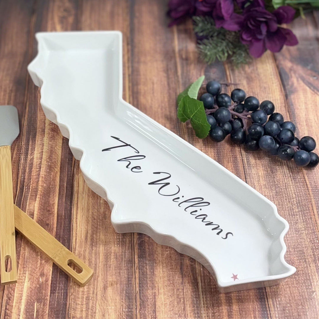 White ceramic tray shaped like a state with 'The Williams' imprinted, on a wooden surface with grapes and a spatula.