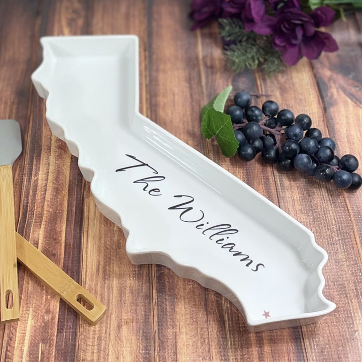 White ceramic tray shaped like a state with 'The Williams' imprinted, on a wooden surface with grapes and a spatula.