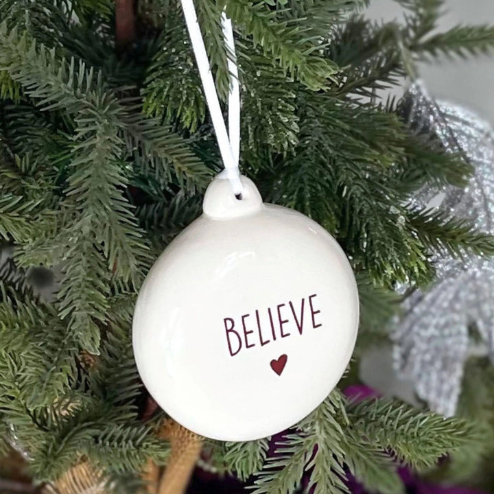 A glossy white ceramic Christmas ornament hanging from a green Christmas tree branch. The ornament features the word 'BELIEVE' in sepia lettering above a small heart, with a white hanging string attached at the top.