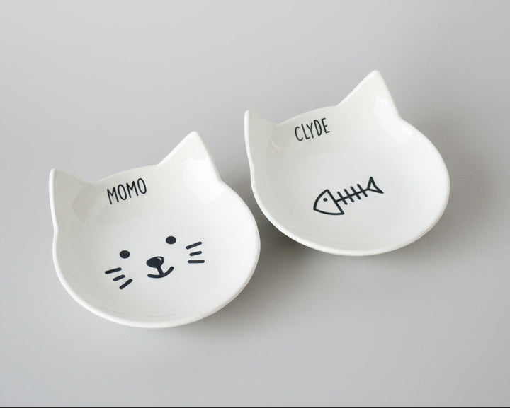 Two white ceramic cat-shaped dishes labeled personalized with your cat's names.
