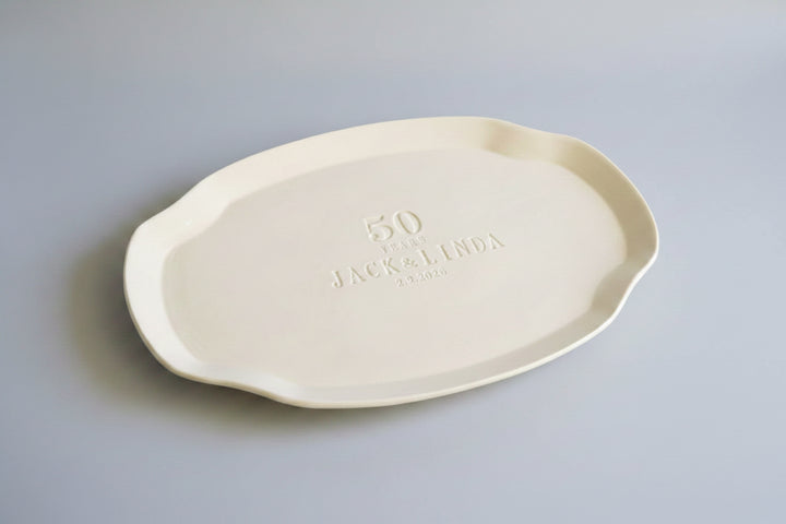 White ceramic platter with '50' and 'Jack & Linda' text, accompanied by flowers on a dark surface.