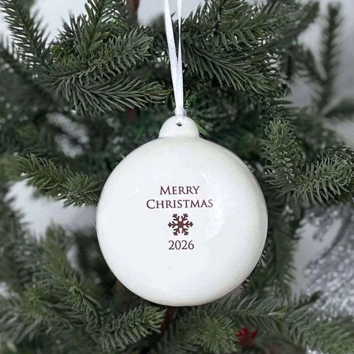 A white spherical Christmas ornament hanging from a green evergreen tree branch, printed with 'MERRY CHRISTMAS 2026' and a small snowflake design.
