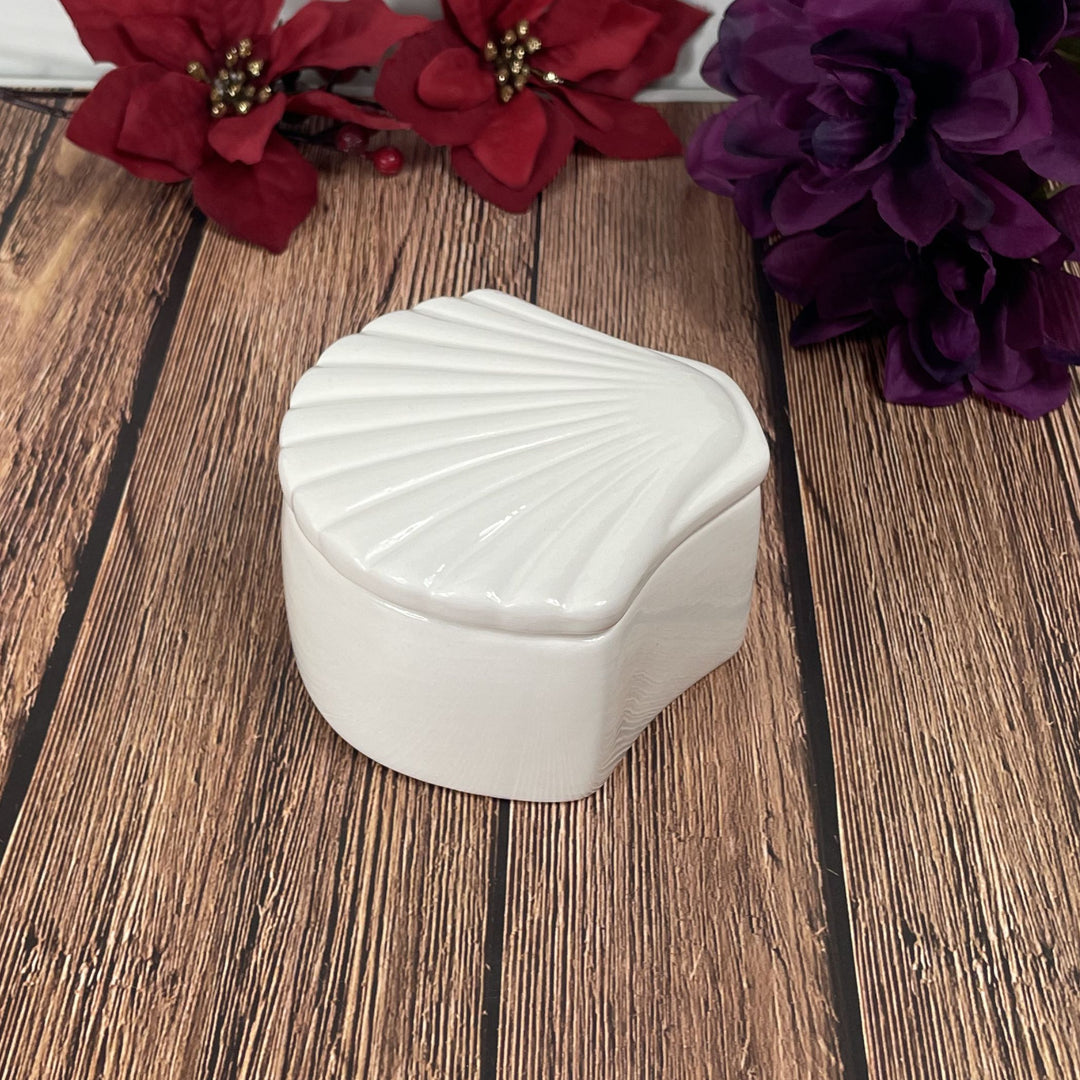 White shell-shaped container on a wooden surface with red and purple flowers in the background
