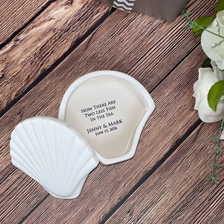 Two white shell-shaped coasters with engraved text on a wooden surface, accompanied by white roses.