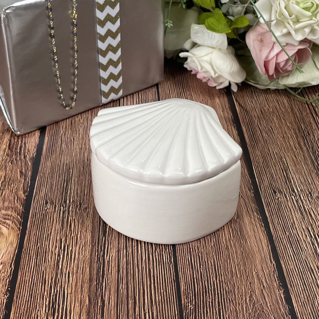 White shell-shaped jewelry box on a wooden surface with flowers and a gift box in the background.