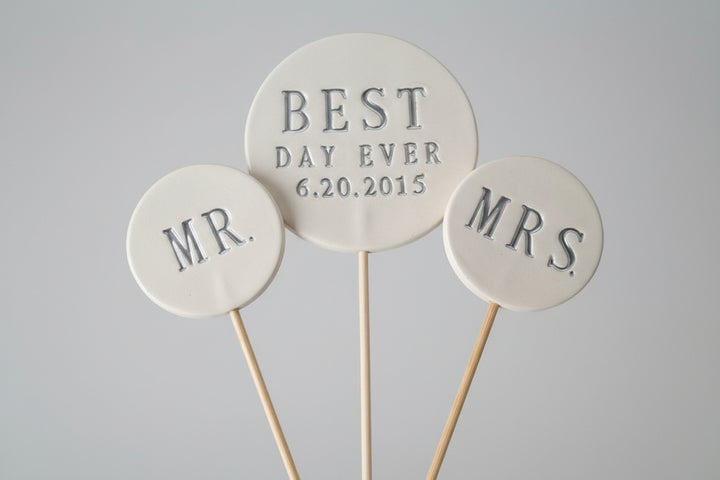 Three round wedding toppers labeled 'MR', 'BEST DAY EVER 6.20.2015', and 'MRS' on a wooden surface.