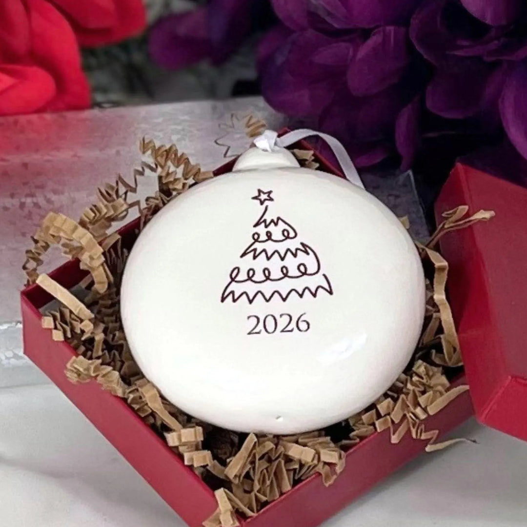 A white round Christmas ornament with a simple tree design and the year 2026, presented in a red gift box with brown crinkle paper filler. Red flowers are blurred in the background.